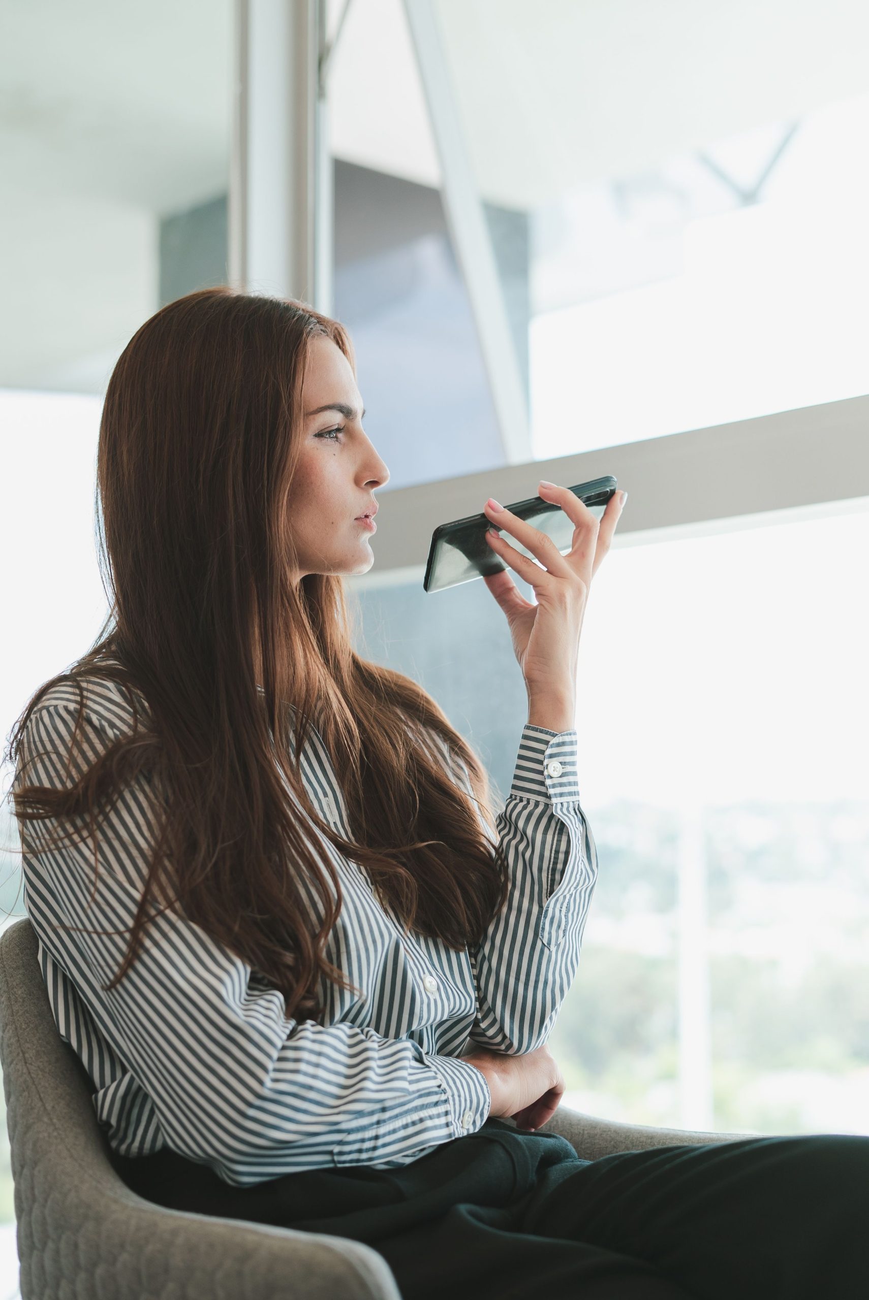 Young businesswoman using voice assistant on her phone in a modern office setting.